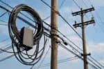 Telecomunicações: Postes de madeira com fios elétricos e de telecomunicações, incluindo uma caixa de distribuição de fibra óptica, sob um céu parcialmente nublado.